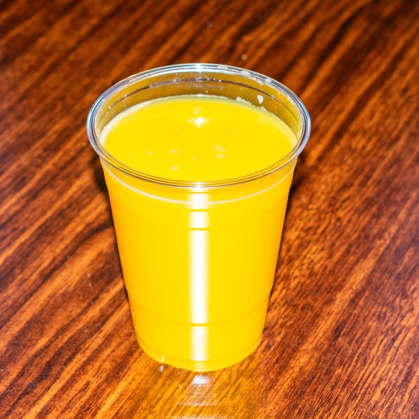 A clear plastic cup filled with bright yellow orange juice sits on a wooden table.