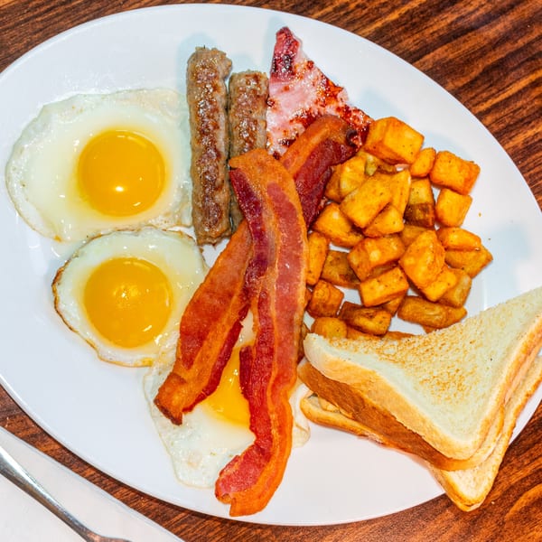 A full breakfast plate with fried eggs, bacon, sausage, home fries, and toast on a white plate.