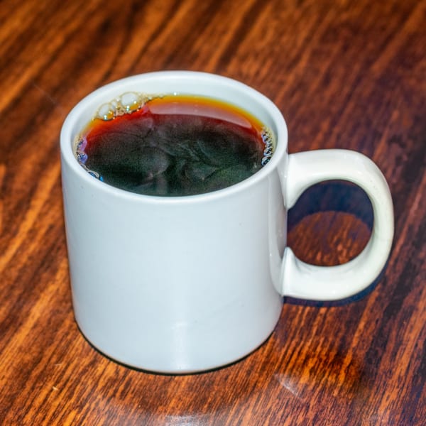 A white mug filled with dark, steaming coffee sits on a rich brown wooden table.