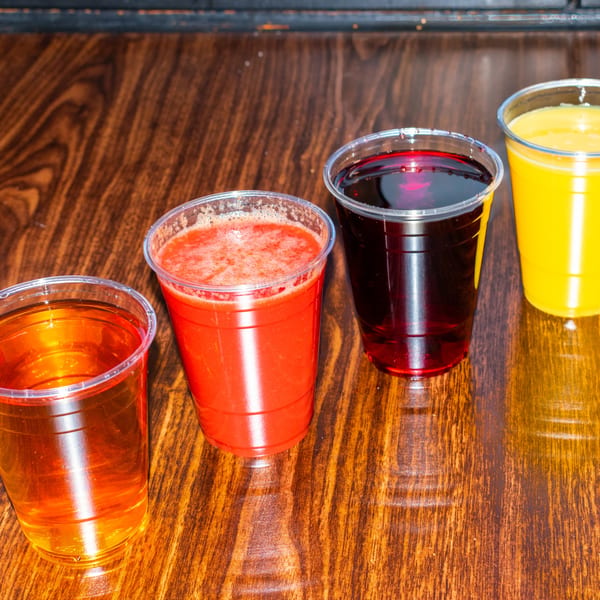 Four plastic cups of J's Brothers Restaurant's colorful juices and smoothies on a wooden table.